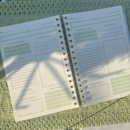 Open spiral-bound planner with various sections on a textured green surface