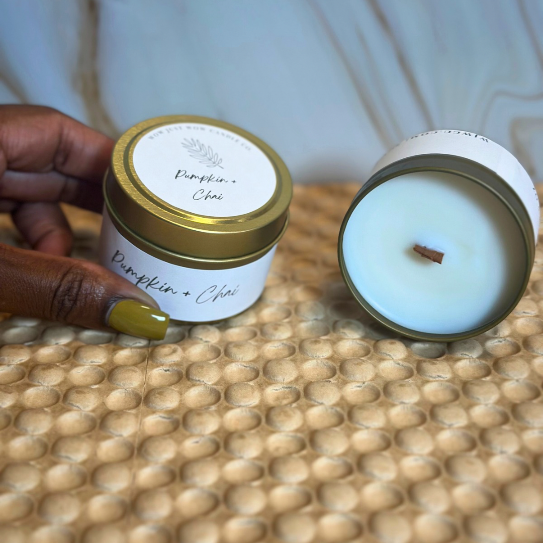 Two soy candles: gold-lidded Pumpkin + Chai tin and blue tin with unlit wick, hand-held.