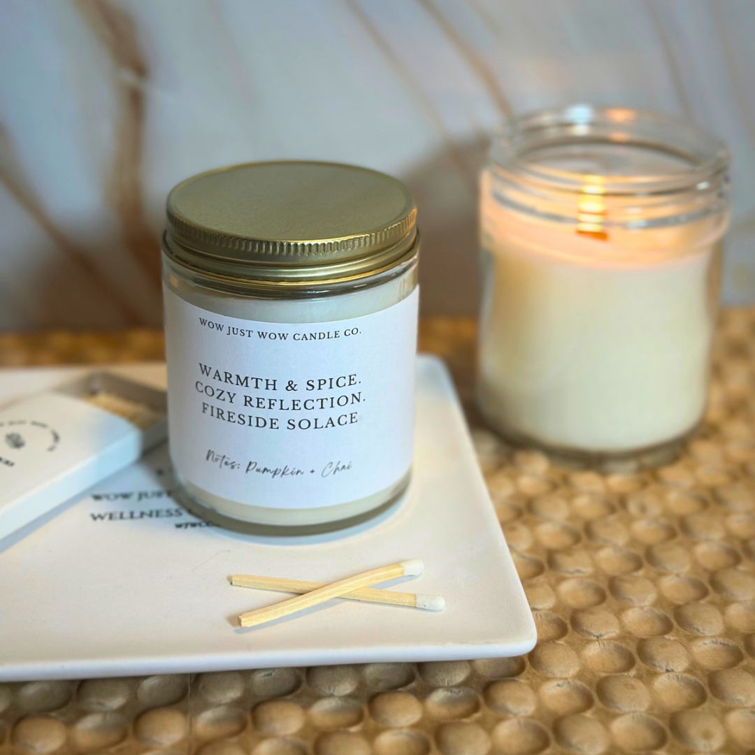 Candle jar with a gold lid on a coaster next to a lit candle on a textured surface.
