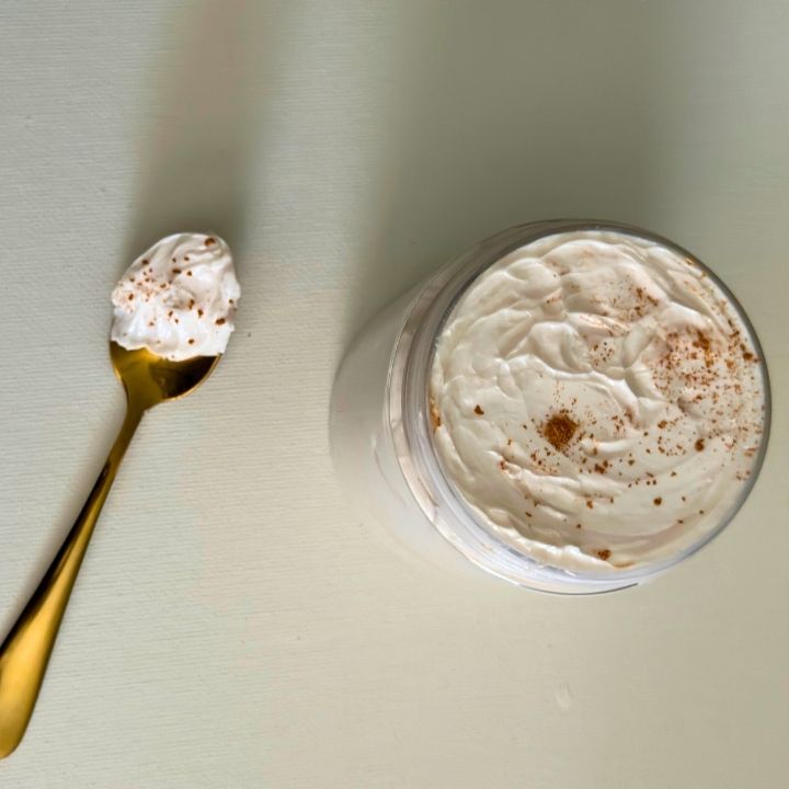 A jar of creamy whipped body butter next to a gold-colored spoon, both with a white creamy product on them, on a light background.